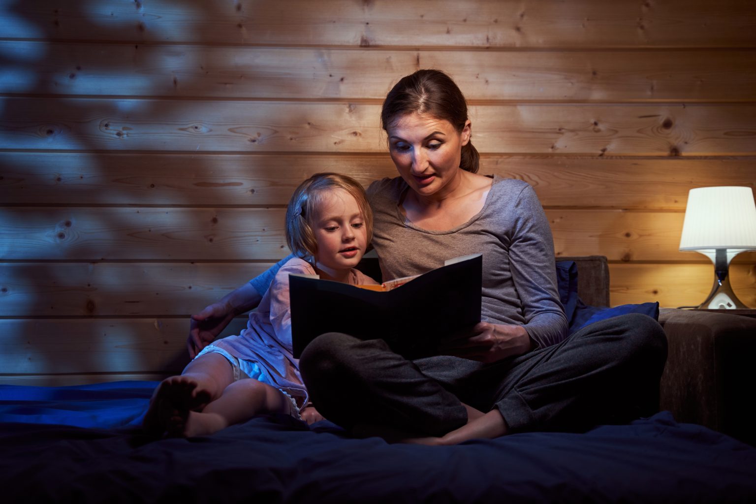 Woman sitting with a child on bed with a night lamp nearby and reading her a book with fairy tales
